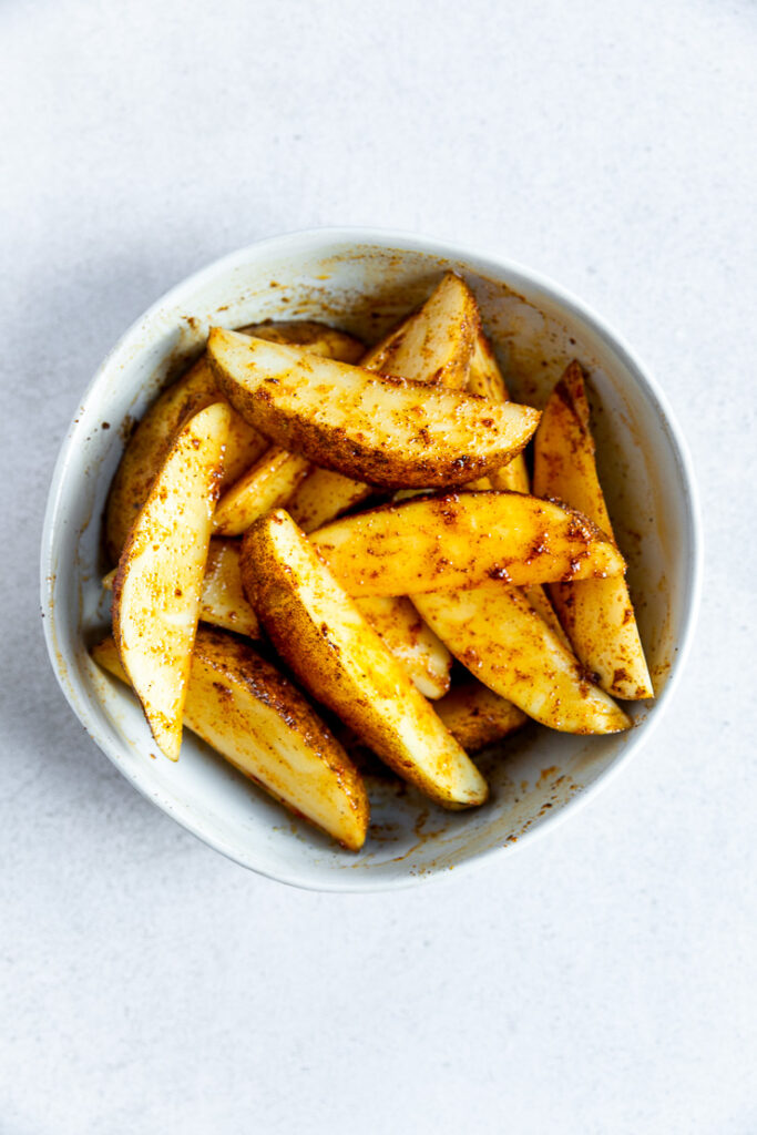 A white bowl filled with raw potato wedges covered in olive oil and seasoning.