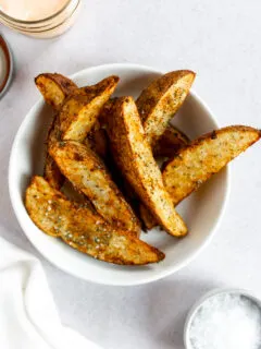 Crispy air fryer potato wedges sitting on top of each other in a white bowl with flaky salt on top.