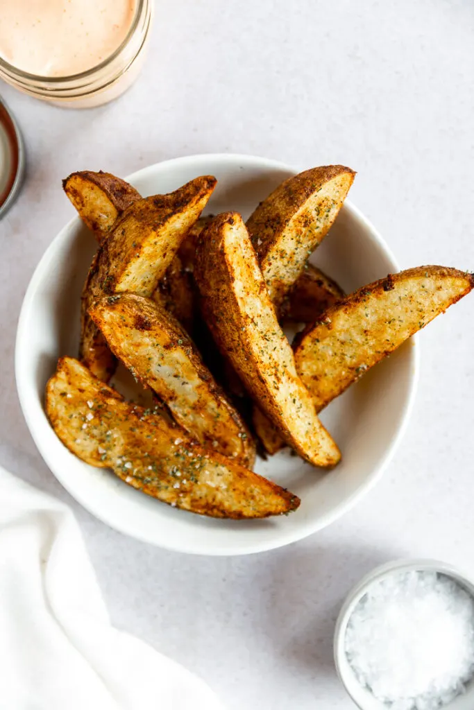 Crispy air fryer potato wedges sitting on top of each other in a white bowl with flaky salt on top.