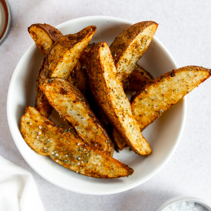 Crispy air fryer potato wedges sitting on top of each other in a white bowl with flaky salt on top.