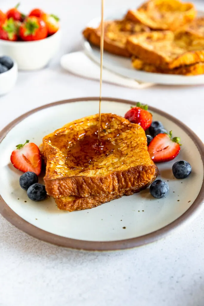 Maple syrup being poured over two pieces of brioche french toast next to berries on a plate.
