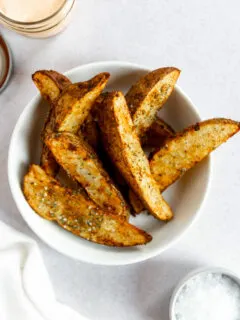 Crispy air fryer potato wedges sitting on top of each other in a white bowl with flaky salt on top.