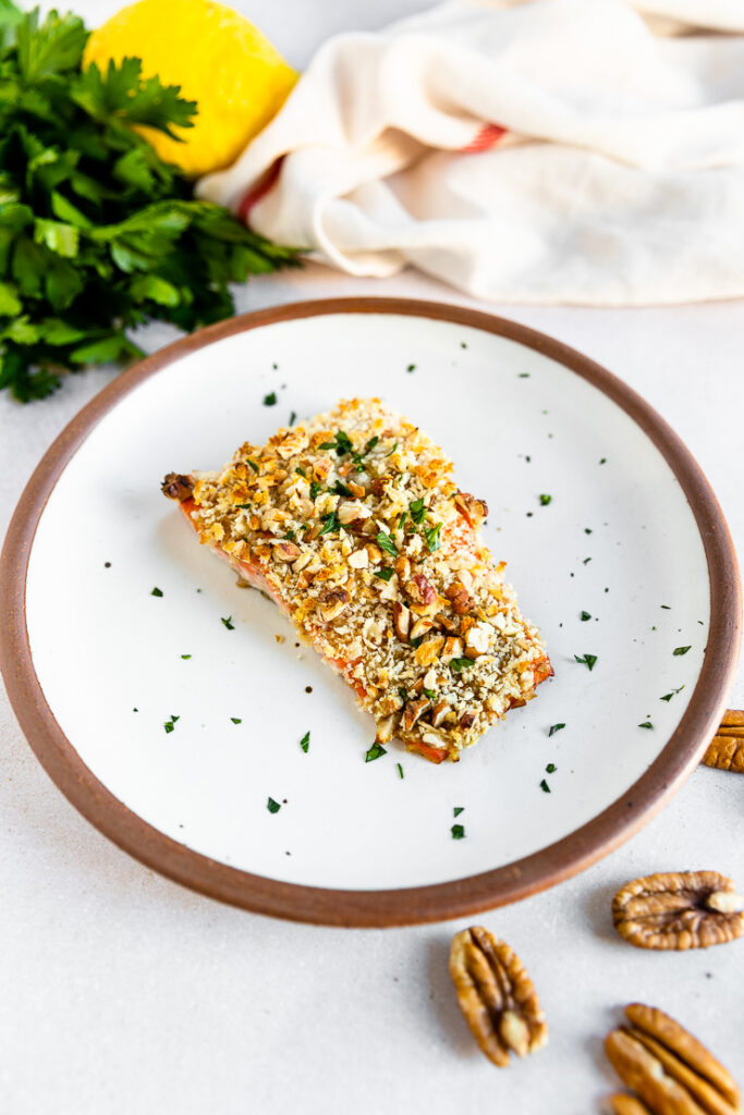 A side shot of a salmon filet topped the pecan breadcrumb mixture and freshly diced parsley on a white plate.