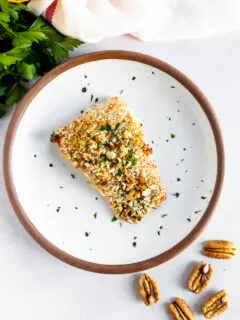 An overhead shot of a salmon filet topped the pecan breadcrumb mixture and freshly diced parsley on a white plate with parsley, lemon, and whole pecans scattered around the plate.