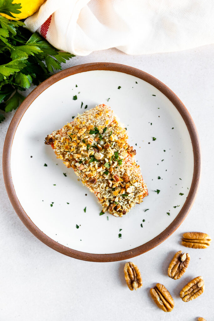 An overhead shot of a salmon filet topped the pecan breadcrumb mixture and freshly diced parsley on a white plate with parsley, lemon, and whole pecans scattered around the plate.