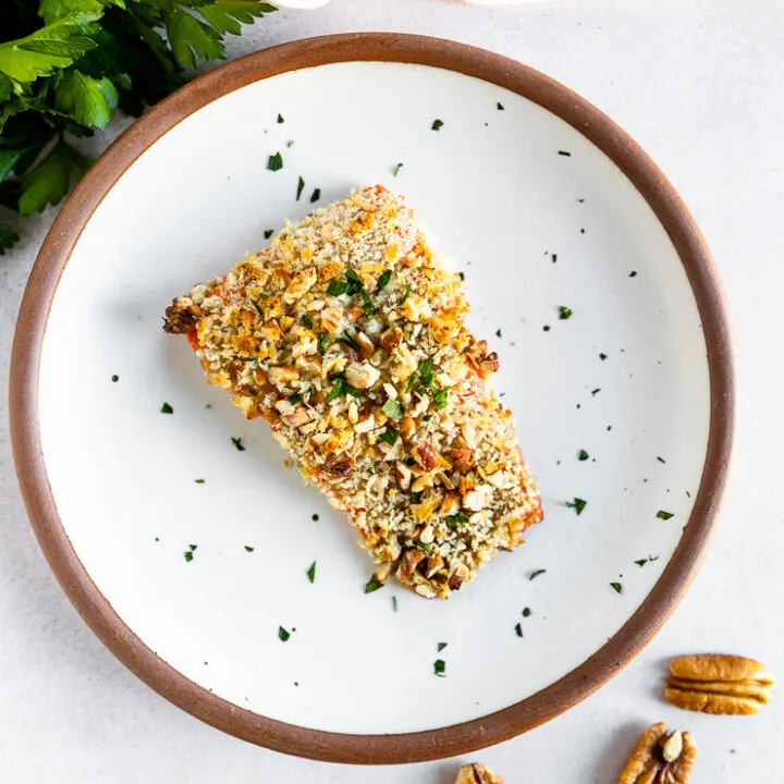 An overhead shot of a salmon filet topped the pecan breadcrumb mixture and freshly diced parsley on a white plate with parsley, lemon, and whole pecans scattered around the plate.