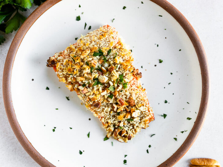 An overhead shot of a salmon filet topped the pecan breadcrumb mixture and freshly diced parsley on a white plate with parsley, lemon, and whole pecans scattered around the plate.