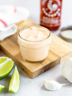 A small mason jar filled with sriracha aioli on a cutting board surrounded by ingredients.