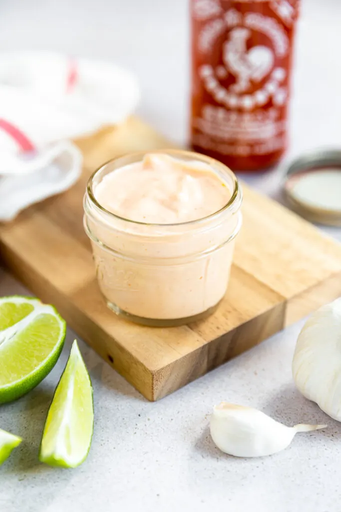 A small mason jar filled with sriracha aioli on a cutting board surrounded by ingredients.