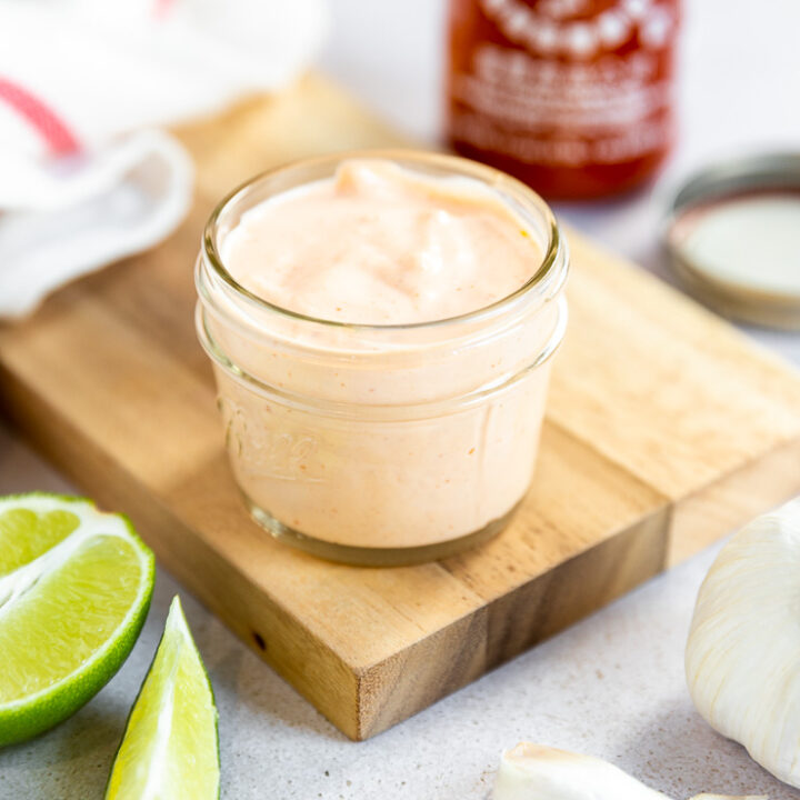 A small mason jar filled with sriracha aioli on a cutting board surrounded by ingredients.