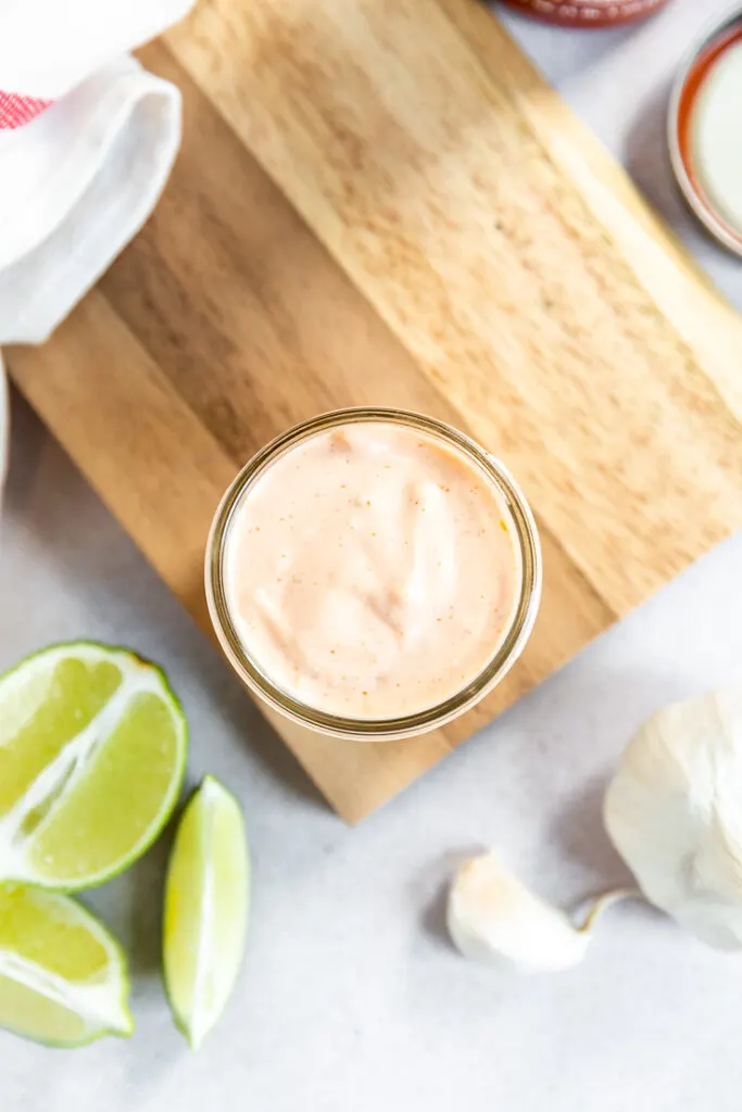 An overhead shot of a the mason jar filled with sriracha aioli next to a half of lime and garlic head.