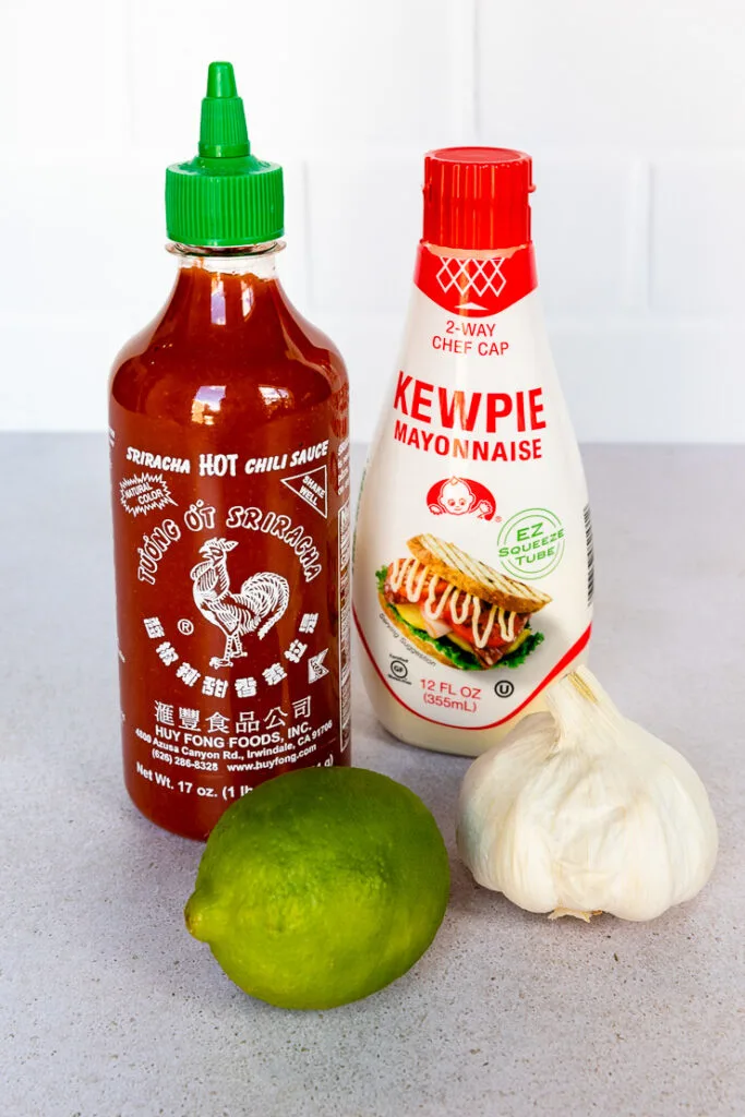 A bottle of Sriracha, Kewpie Mayonnaise, a lime, and a head of garlic all sitting next to each other.