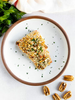 An overhead shot of a salmon filet topped the pecan breadcrumb mixture and freshly diced parsley on a white plate with parsley, lemon, and whole pecans scattered around the plate.