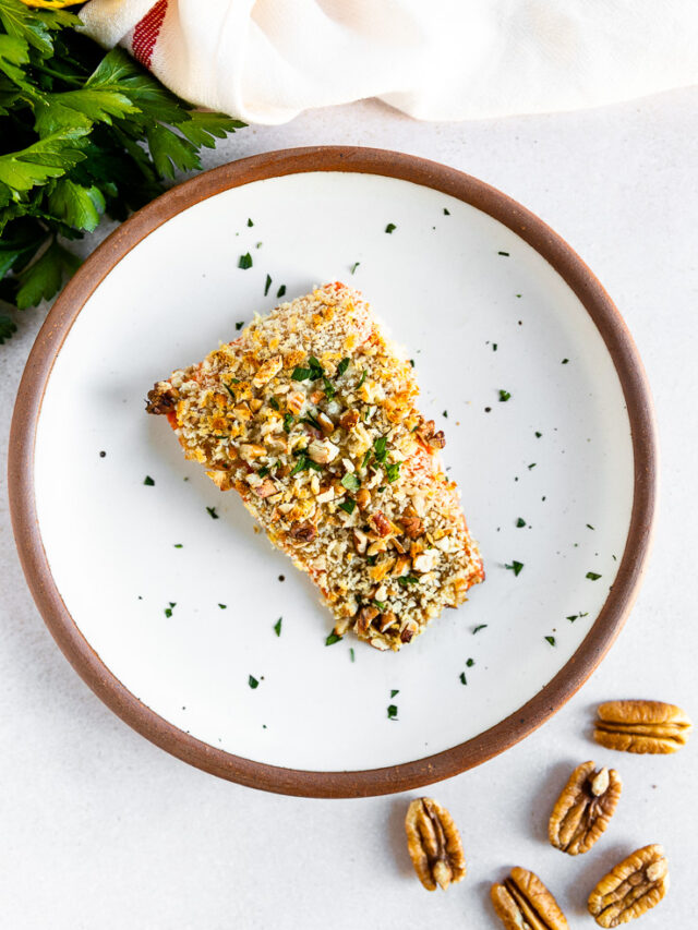 An overhead shot of a salmon filet topped the pecan breadcrumb mixture and freshly diced parsley on a white plate with parsley, lemon, and whole pecans scattered around the plate.