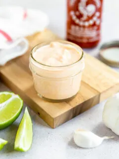 A small mason jar filled with sriracha aioli on a cutting board surrounded by ingredients.