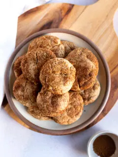 An overhead shot looking at a plate of cookies stacked high with spice cake mix cookies on a wooden board next to a small bowl with leftover cinnamon sugar mixture.