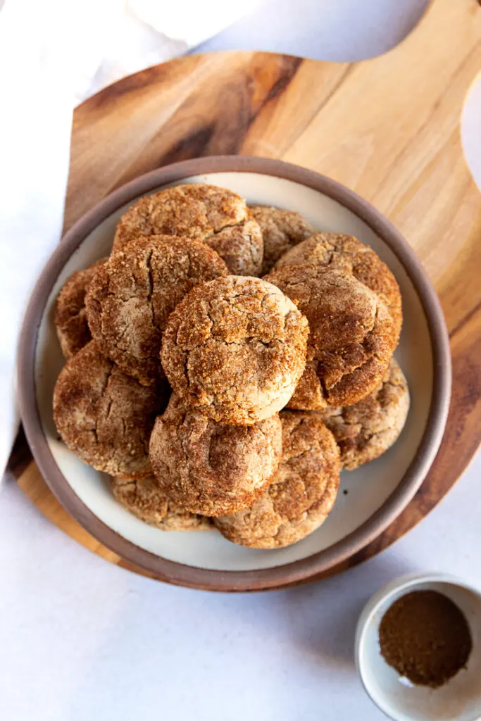 An overhead shot looking at a plate of cookies stacked high with spice cake mix cookies on a wooden board next to a small bowl with leftover cinnamon sugar mixture.