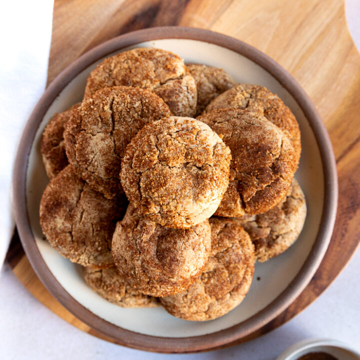 An overhead shot looking at a plate of cookies stacked high with spice cake mix cookies on a wooden board next to a small bowl with leftover cinnamon sugar mixture.