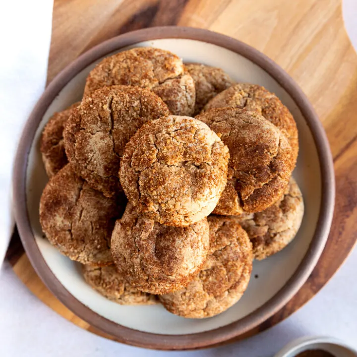 An overhead shot looking at a plate of cookies stacked high with spice cake mix cookies on a wooden board next to a small bowl with leftover cinnamon sugar mixture.