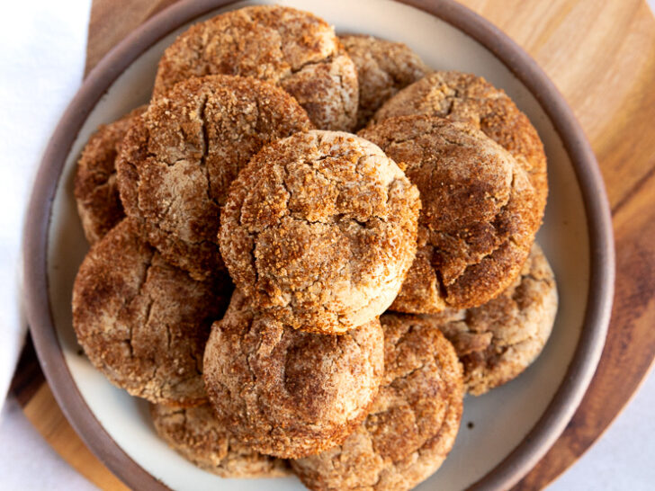 An overhead shot looking at a plate of cookies stacked high with spice cake mix cookies on a wooden board next to a small bowl with leftover cinnamon sugar mixture.