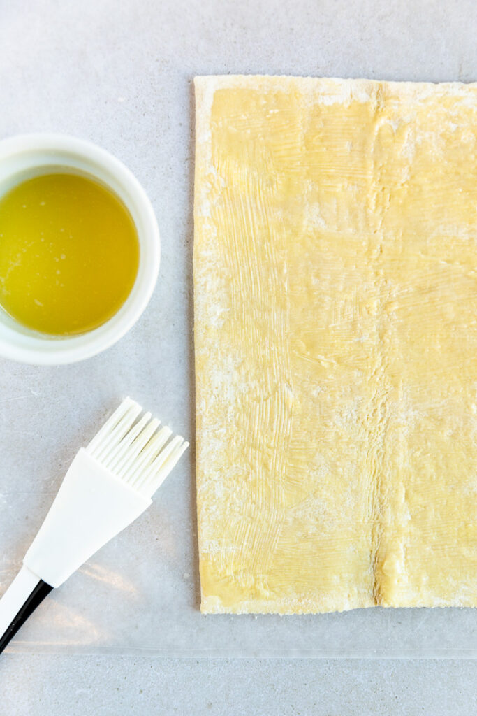 A sheet of puff pastry with melted butter spread evenly on top next to a silicone brush and melted butter in a small bowl.