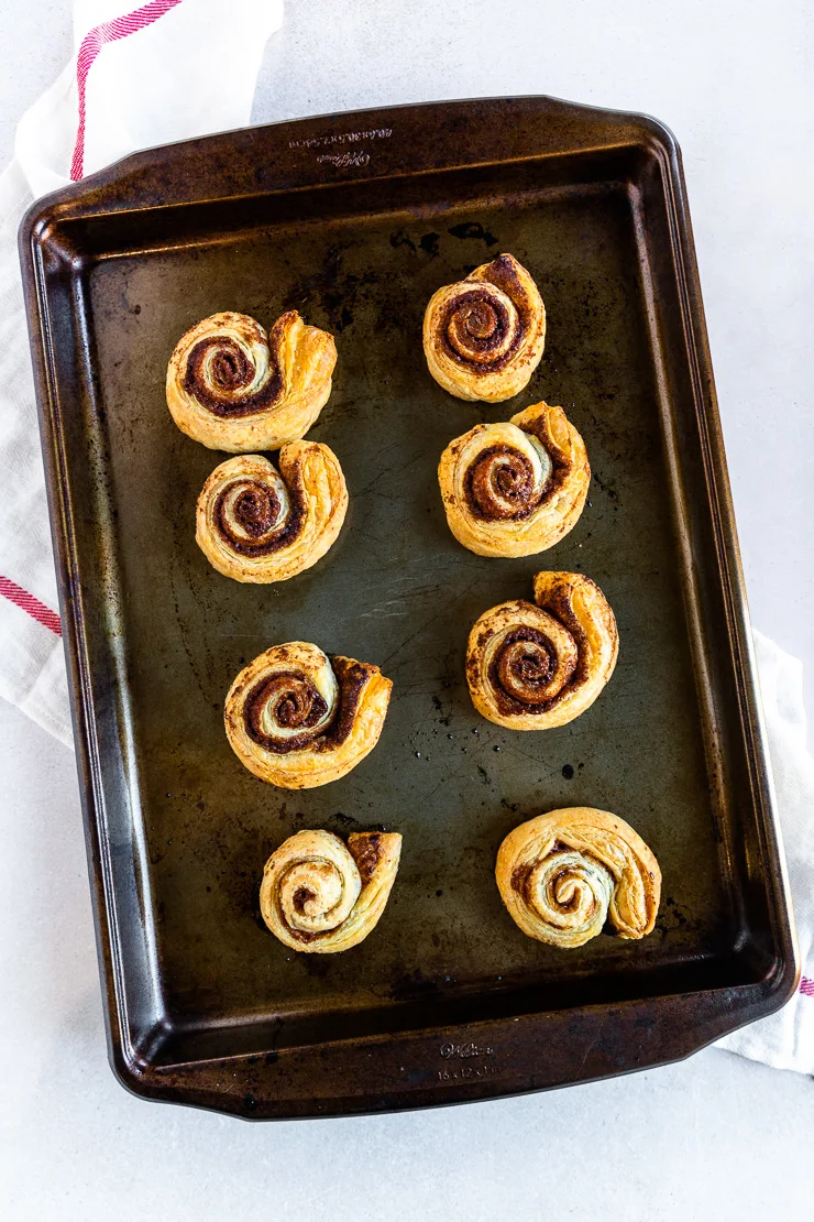 A cookie sheet of eight golden brown puff pastry cinnamon rolls on a cloth napkin.