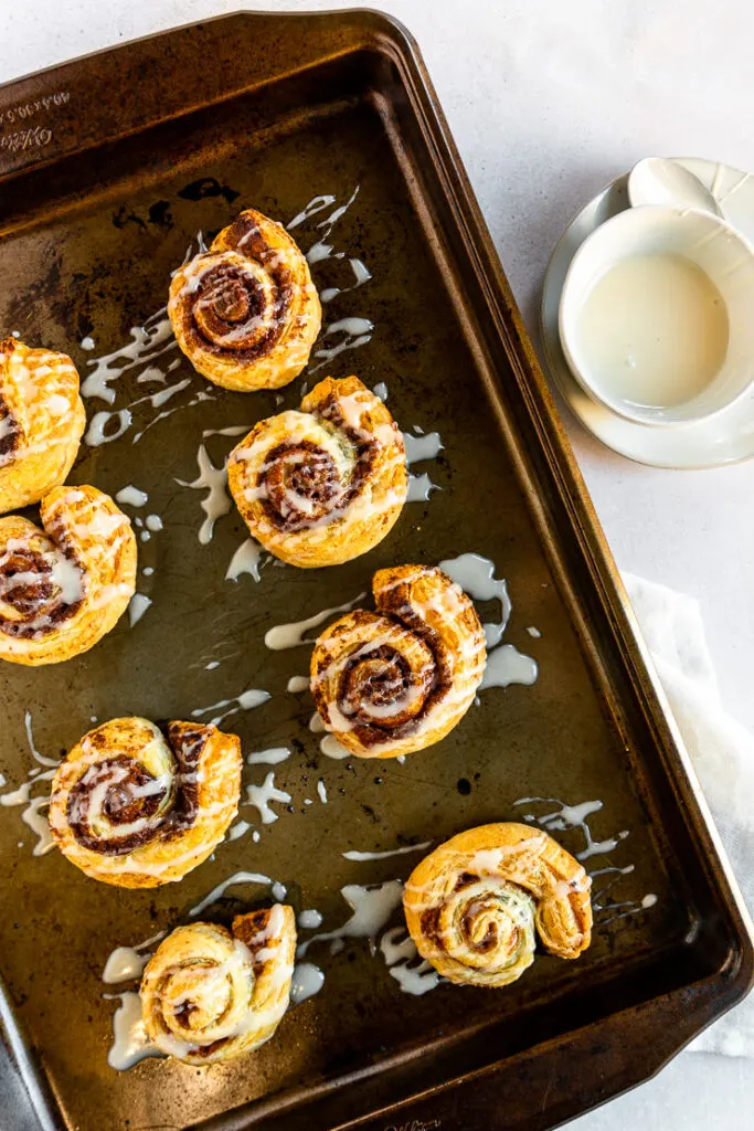 A white sugary topping is drizzled over each of the baked cinnamon rolls on the cookie sheet.