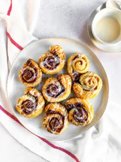 An overhead shot of prepared puff pastry cinnamon rolls on a white plate next to a small bowl of sugary topping.