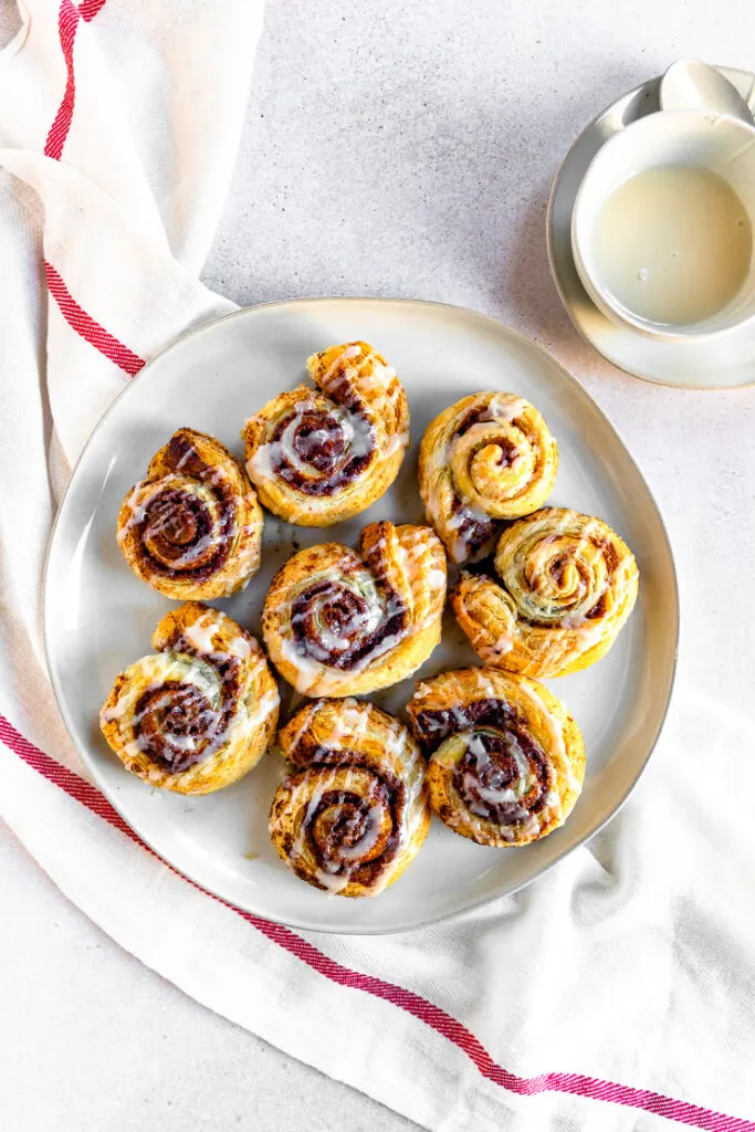 An overhead shot of prepared puff pastry cinnamon rolls on a white plate next to a small bowl of sugary topping.