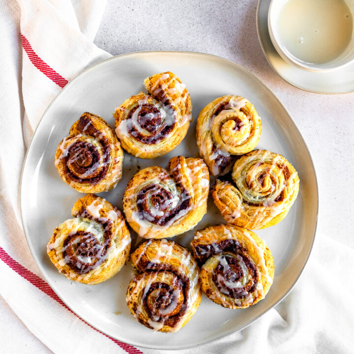 An overhead shot of prepared puff pastry cinnamon rolls on a white plate next to a small bowl of sugary topping.