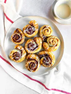 An overhead shot of prepared puff pastry cinnamon rolls on a white plate next to a small bowl of sugary topping.