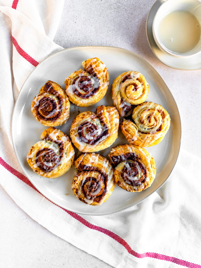 An overhead shot of prepared puff pastry cinnamon rolls on a white plate next to a small bowl of sugary topping.