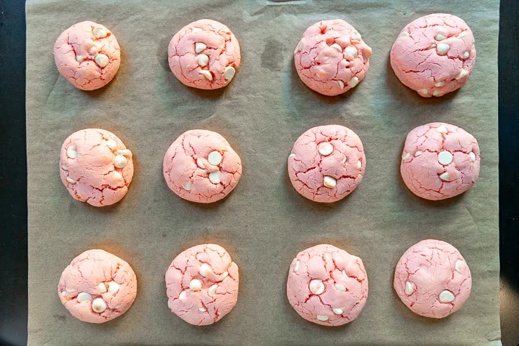 Twelve baked strawberry cake mix cookies with white chocolate chips on a parchment lined baking sheet.