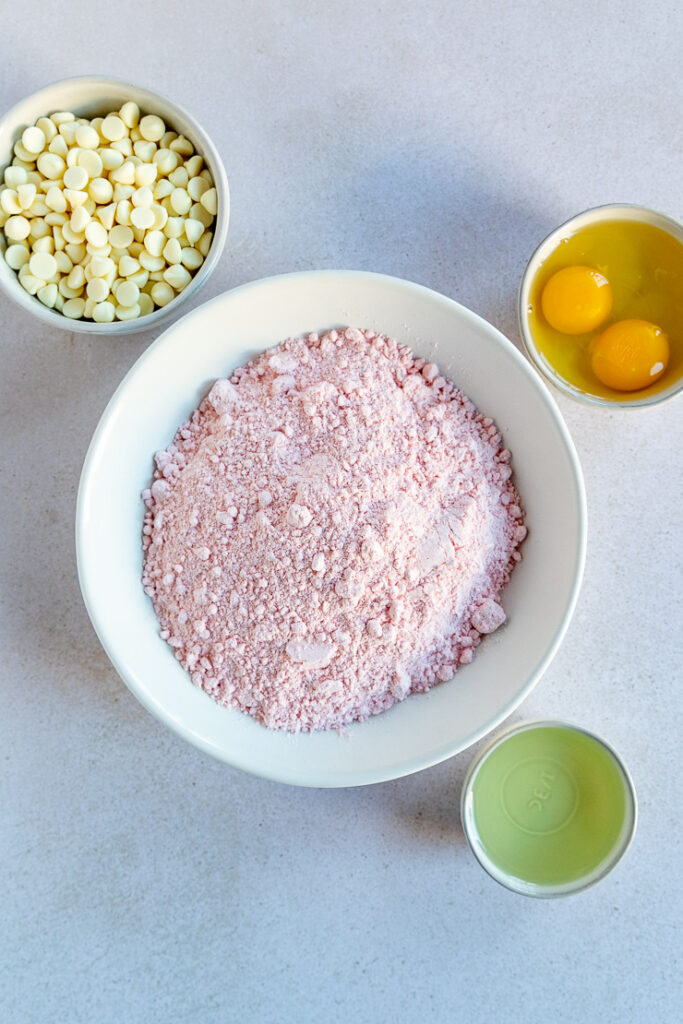 Four white bowls containing the strawberry cake mix, eggs, vegetable oil, and white chocolate chips.