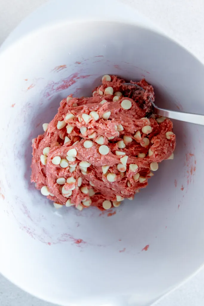 A pink ball of cookie dough with white chocolate chips being stirred with a silver spoon in a white mixing bowl.