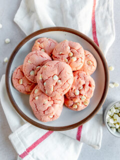 A plate of baked strawberry cake mix cookies on a white cloth napkin with a red stripe next to loose white chocolate chips.