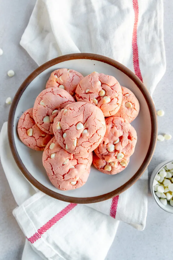 A plate of baked strawberry cake mix cookies on a white cloth napkin with a red stripe next to loose white chocolate chips.