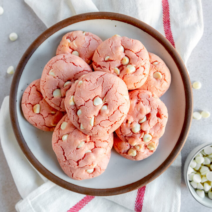 A plate of baked strawberry cake mix cookies on a white cloth napkin with a red stripe next to loose white chocolate chips.