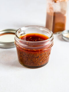 A small glass jar filled with reddish-brown ancho chile marinade sits on a white surface, with a metal lid and an open spice container in the background.