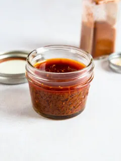 A small glass jar filled with reddish-brown ancho chile marinade sits on a white surface, with a metal lid and an open spice container in the background.