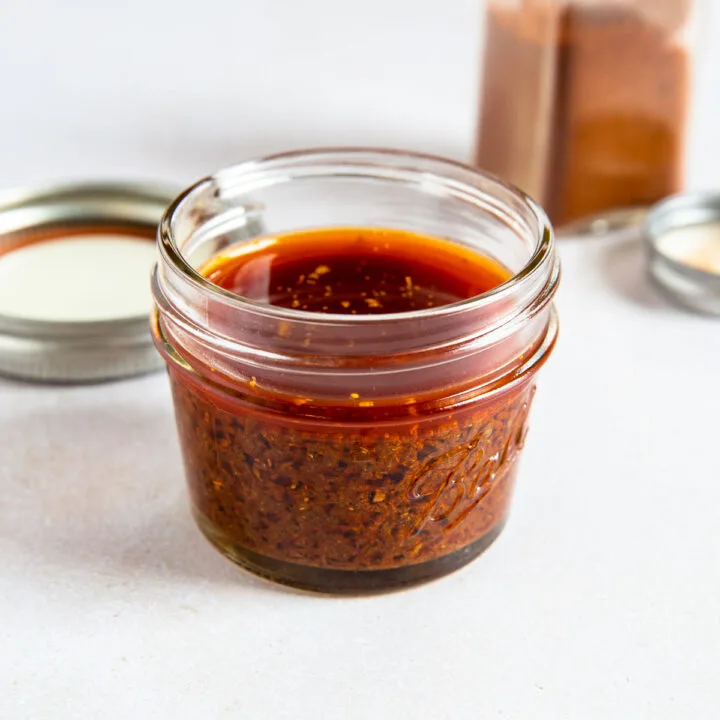 A small glass jar filled with reddish-brown ancho chile marinade sits on a white surface, with a metal lid and an open spice container in the background.