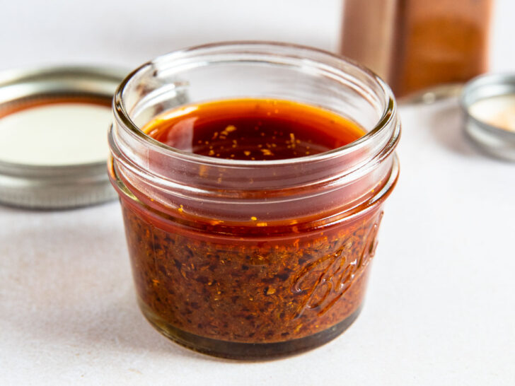 A small glass jar filled with reddish-brown ancho chile marinade sits on a white surface, with a metal lid and an open spice container in the background.