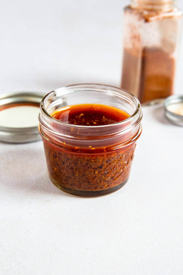 A small glass jar filled with reddish-brown ancho chile marinade sits on a white surface, with a metal lid and an open spice container in the background.