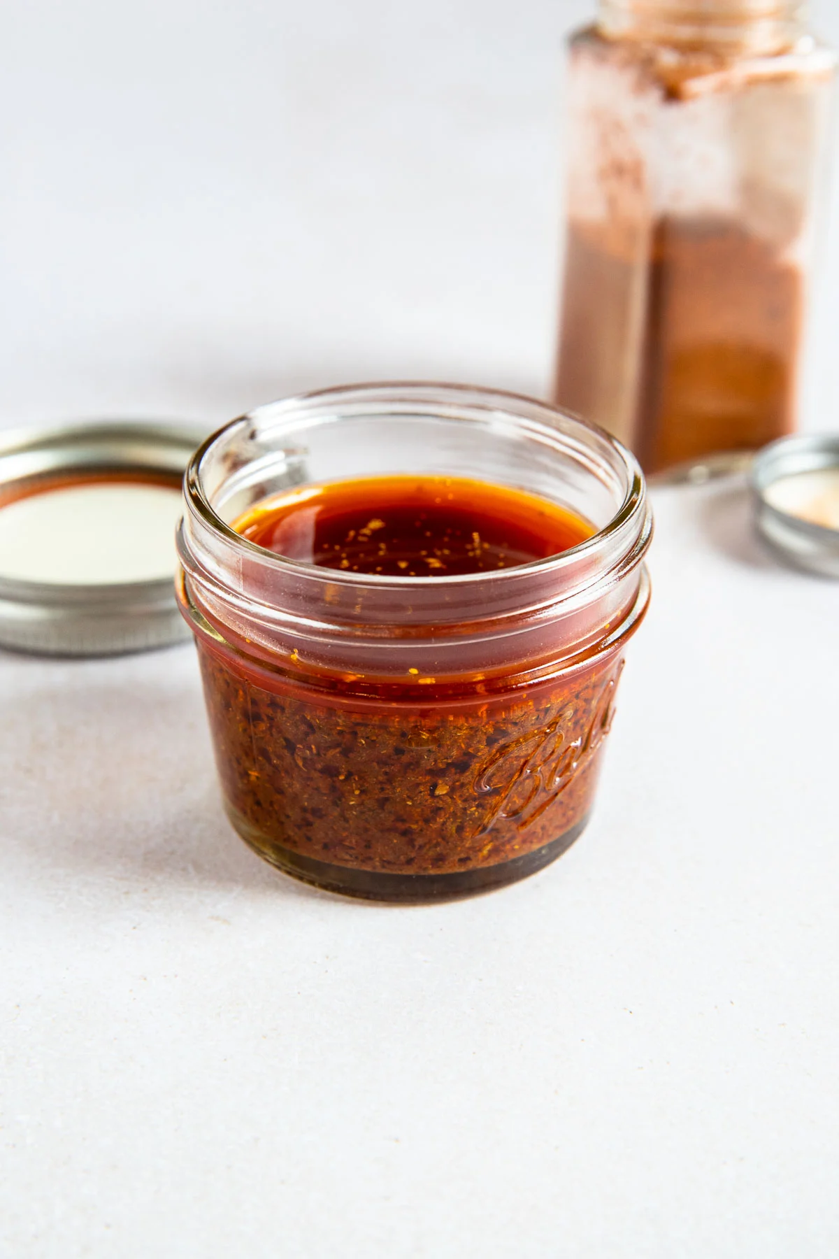 A small glass jar filled with reddish-brown ancho chile marinade sits on a white surface, with a metal lid and an open spice container in the background.