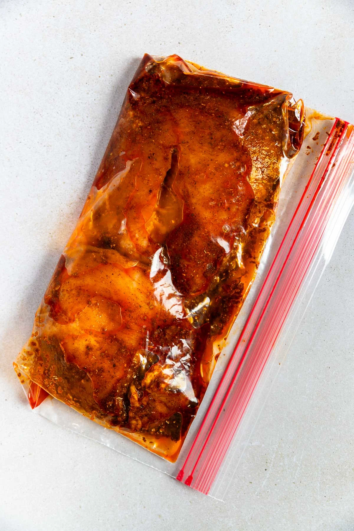 A resealable plastic bag containing pieces of raw chicken marinating in a reddish-brown ancho chicken marinade, placed on a light-colored surface.