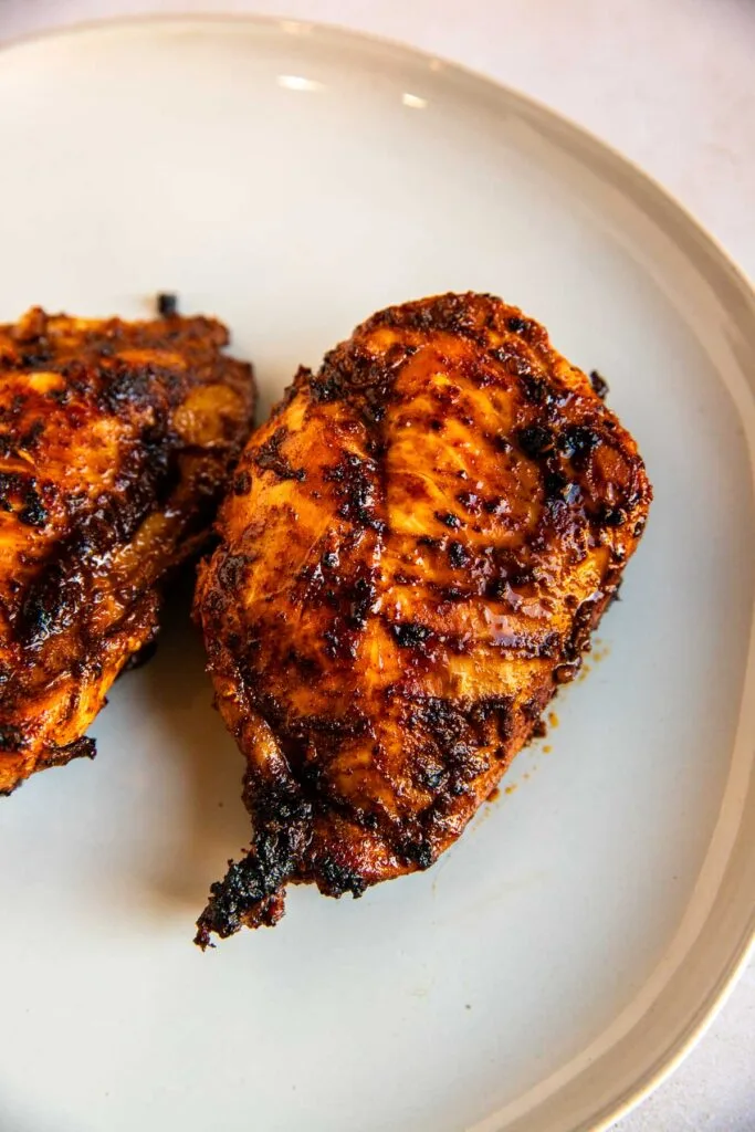 Two pieces of seasoned, grilled chicken breast with a charred, crispy exterior—thanks to a zesty ancho chicken marinade—sit on a plain white plate.
