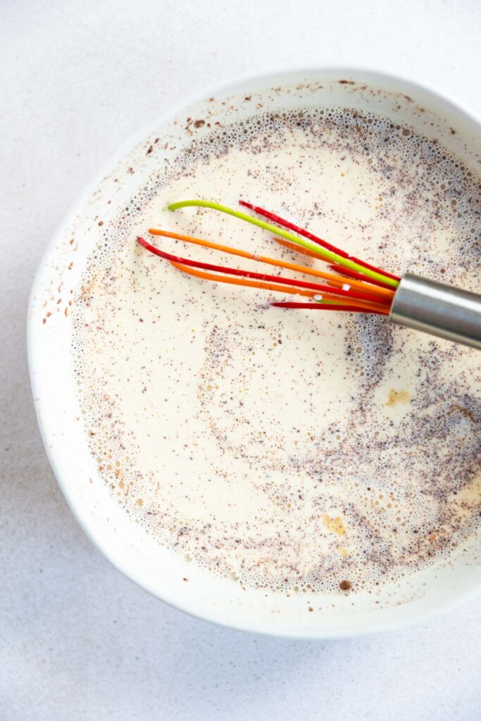 A colorful whisk in a white bowl mixes a creamy liquid with visible specks of spices, perfect for your next brioche French toast recipe.