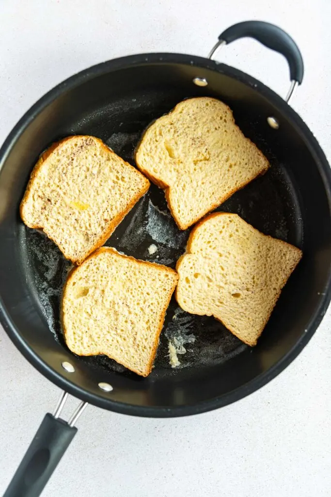 Four slices of bread are toasting in a black nonstick frying pan, perfect for a simple brioche French toast recipe. A small amount of melted butter is visible beneath the bread, and the pan rests on a light-colored countertop.