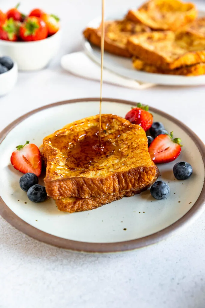 Two slices of french toast with brioche bread on a plate topped with blueberries and halved strawberries, as syrup is being poured over them. A bowl of strawberries and a plate with more french toast are in the background.