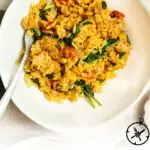 A white plate filled with creamy Italian sausage orzo mixed with spinach and sun-dried tomatoes. A fork rests on the plate. The text below reads: Creamy Italian Sausage Orzo. The Culinary Compass.